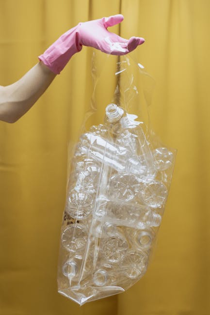 A person wearing a light pink rubber glove is holding a transparent plastic bag filled with crushed and empty plastic bottles, with some bottles visible through the bag's crinkled surface. The backgro