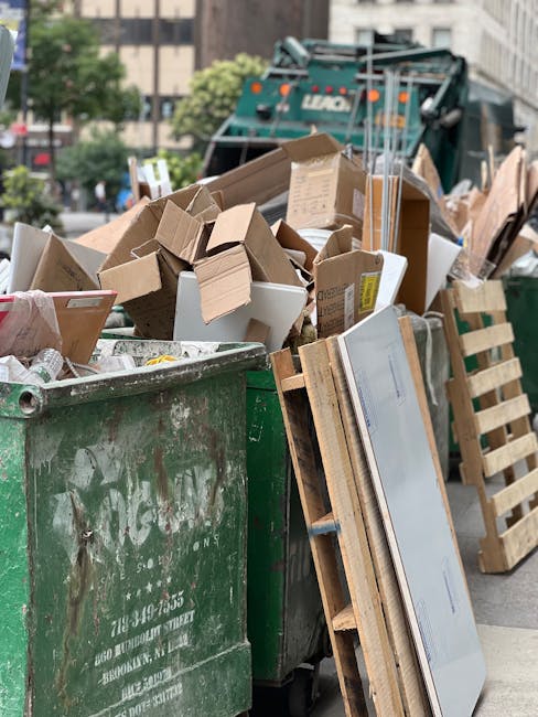A large green waste disposal bin filled with various discarded materials, including numerous cardboard boxes of different sizes and shapes, some flattened and others partially assembled, as well as a 