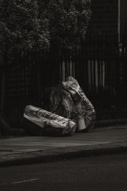 A person wearing light blue jeans and white sneakers is crouching on an asphalt surface, holding a small piece of cluttered debris consisting of styrofoam, tangled cords, and scraps of wood or cardboa