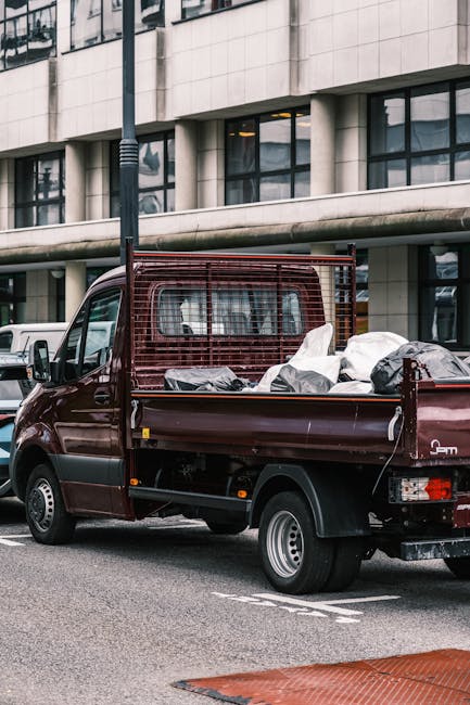 A maroon flatbed utility truck parked on a city street, loaded with various types of waste and rubbish bags, some black and some white, stacked in the truck's open cargo area. The truck has a metal me