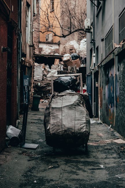 A maroon flatbed utility truck parked on a city street, loaded with various types of waste and rubbish bags, some black and some white, stacked in the truck's open cargo area. The truck has a metal me